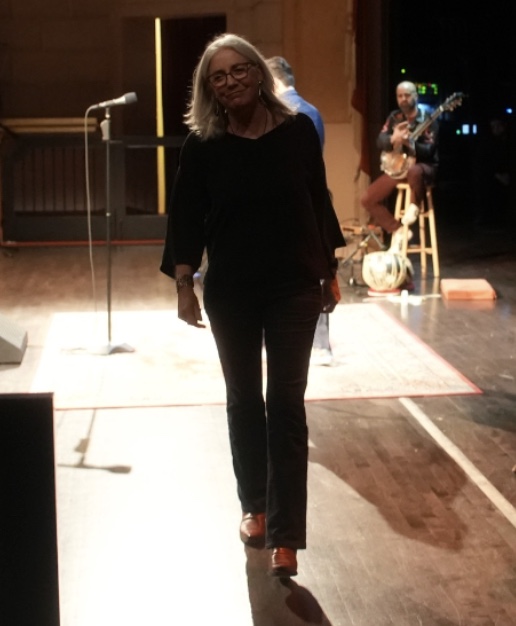 Cathleen walking off stage from telling a story at the Herbst theater. There is a guitar accompaniment in the back corner.