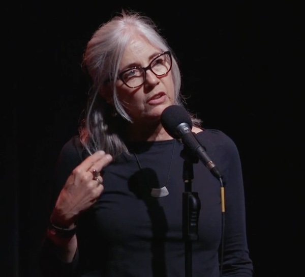 Kathleen speaking in front of a microphone. She has medium-length white hair and large black glasses. She wears a black T-shirt.